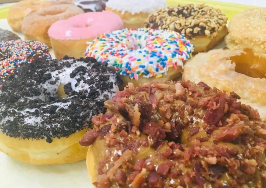 Assorted donuts: maple bacon in front, with Oreo, rainbow sprinkle, strawberry frosted and chocolate peanut donuts behind